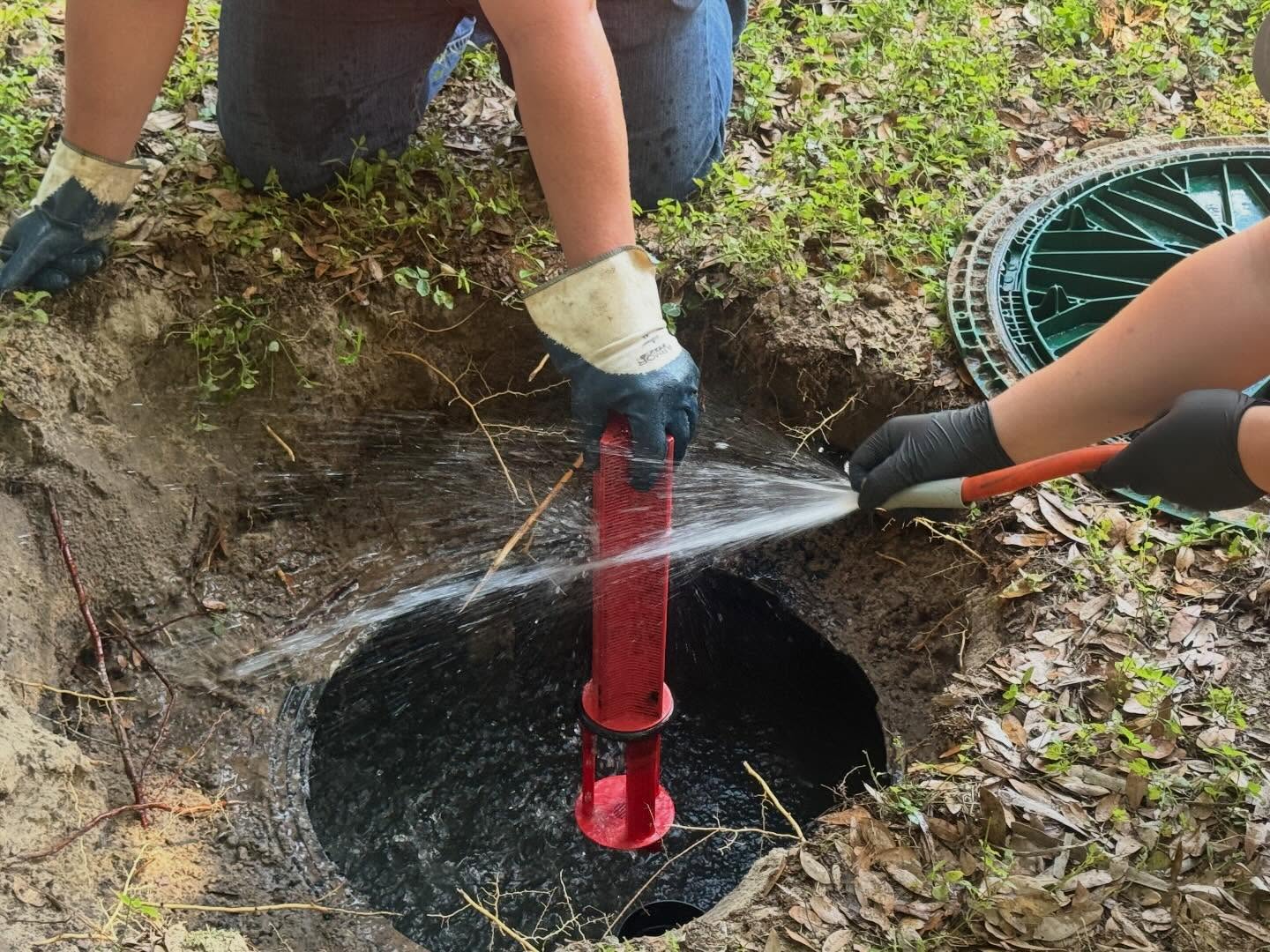 Two septic service technicians perform maintenance on an open septic system, using a hose to clean equipment inside the access opening